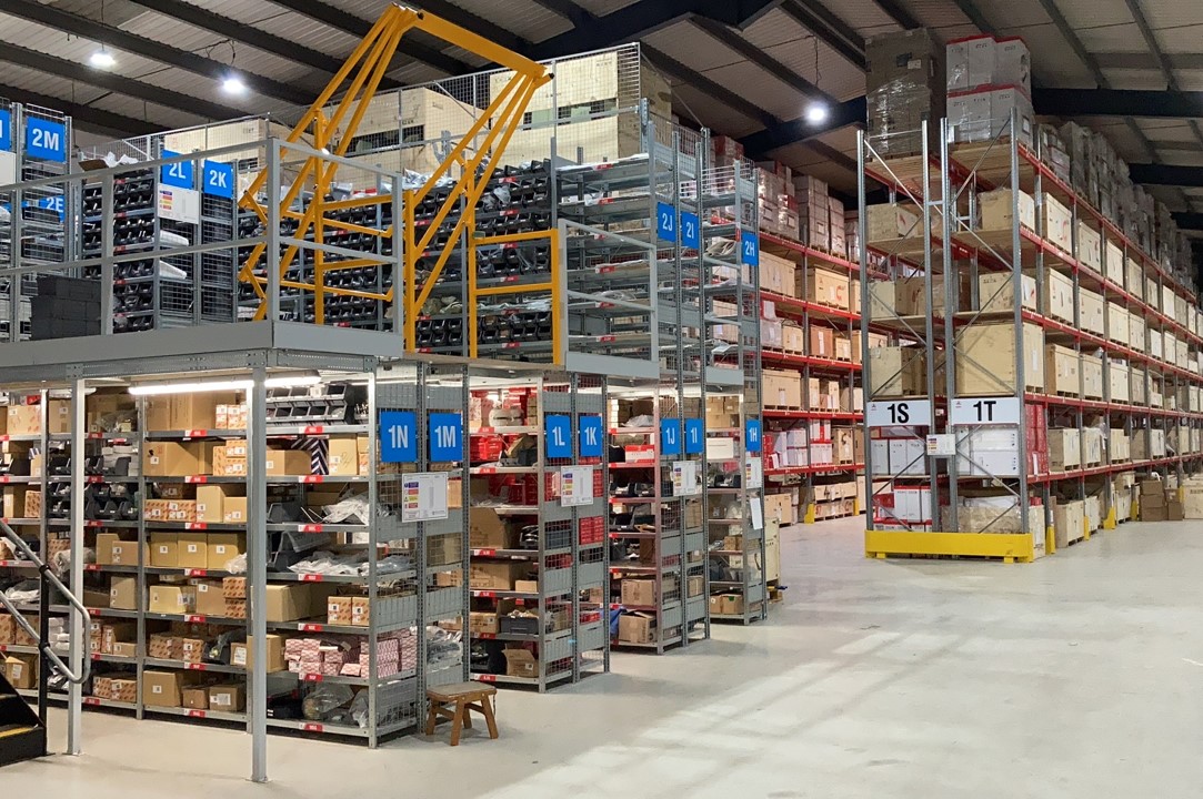 Multi-level auto parts warehouse with pallet racking, labeled storage zones, and boxed inventory arranged by SKU categories for inventory width management
