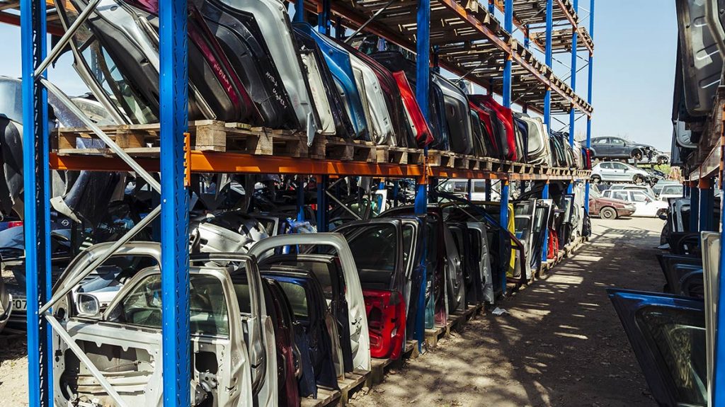 Outdoor automotive body parts storage yard with multiple car doors stacked vertically on steel racks, showing high-volume body panel inventory under open-air conditions.