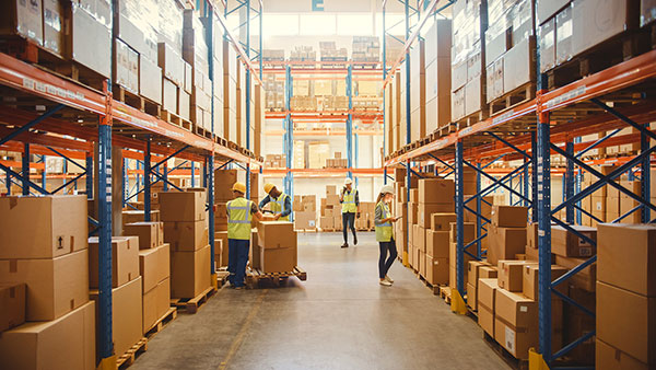 Warehouse aisle with pallet racking filled with boxed inventory, workers handling cartons between shelves and floor storage areas.