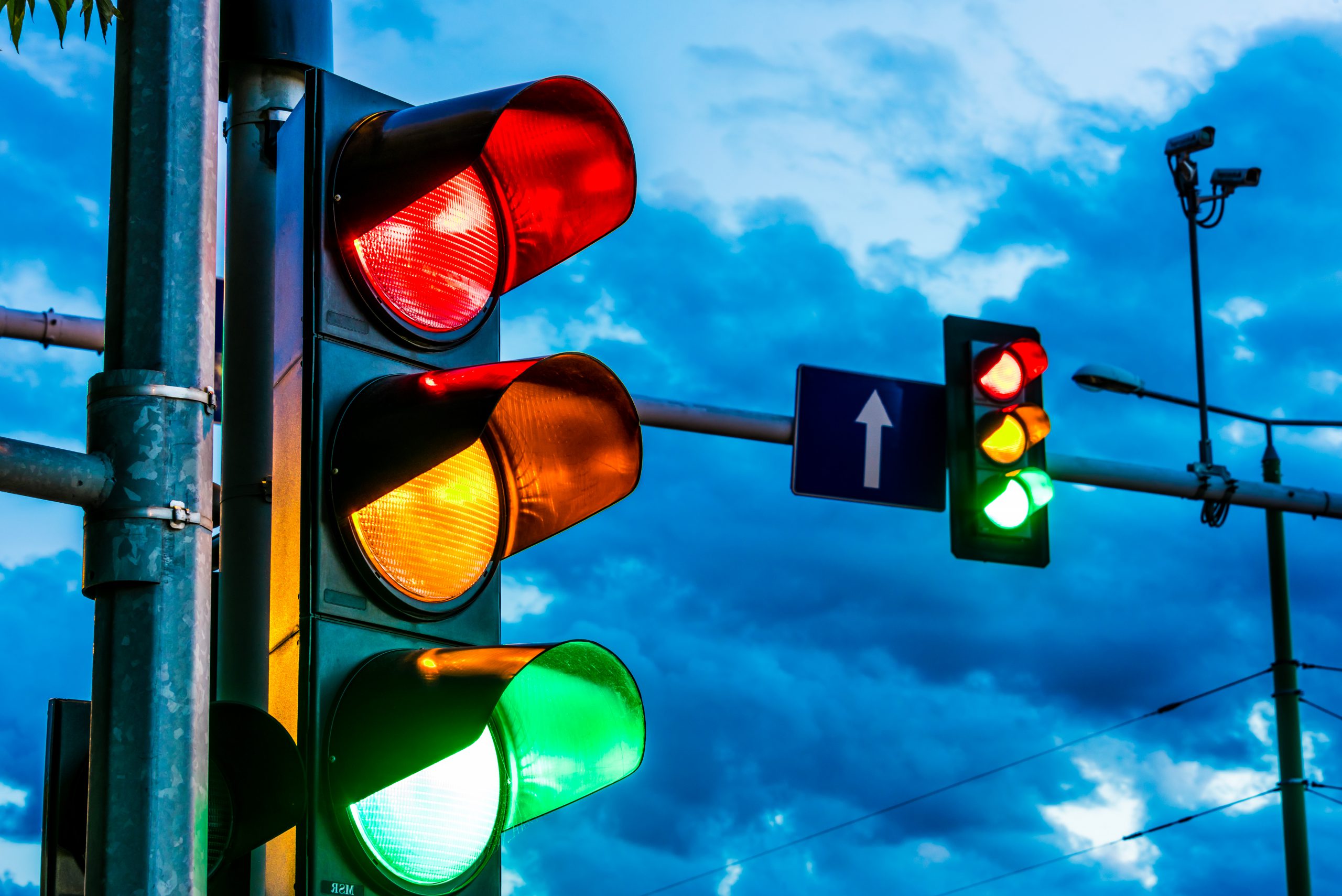 Traffic lights showing red, yellow, and green signals at an intersection under cloudy sky, with directional road signs in background.
