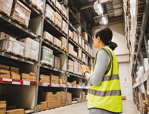 Warehouse worker wearing safety vest checking boxed inventory on high storage shelves